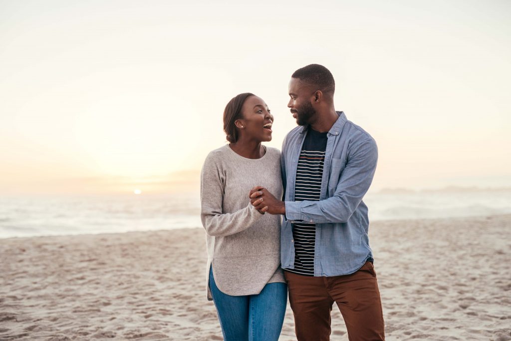 Content young African couple laughing and talking while walking hand in hand together down a sandy beach at dusk