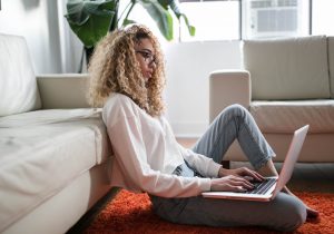 Sitting on the floor, leaning against a couch, a person types on a laptop