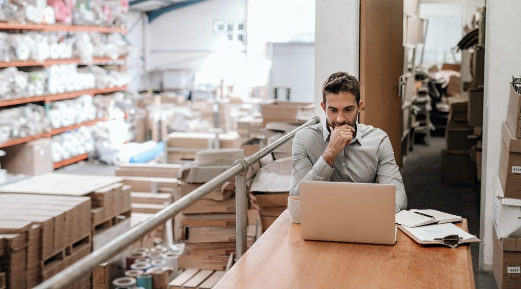 A man sits at a desk with a laptop in a warehouse, surrounded by boxes and shelves of inventory, appearing focused on his work.