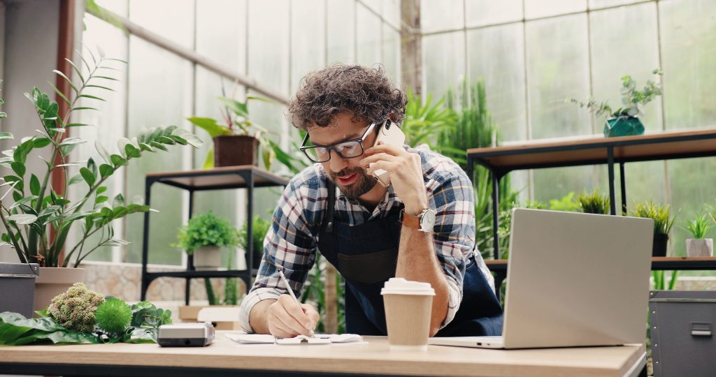 Man wearing glasses and apron talks on phone, writes on paper, and works at a laptop in a plant-filled indoor workspace. Coffee cup and calculator are on the desk.