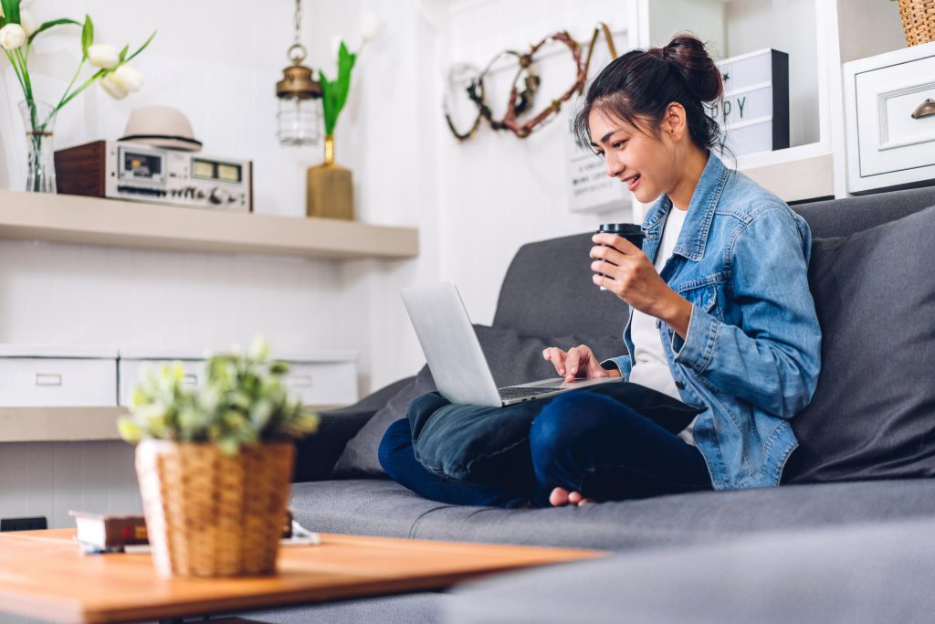 Woman sitting cross-legged on coach with laptop on her lap and coffee in her hand