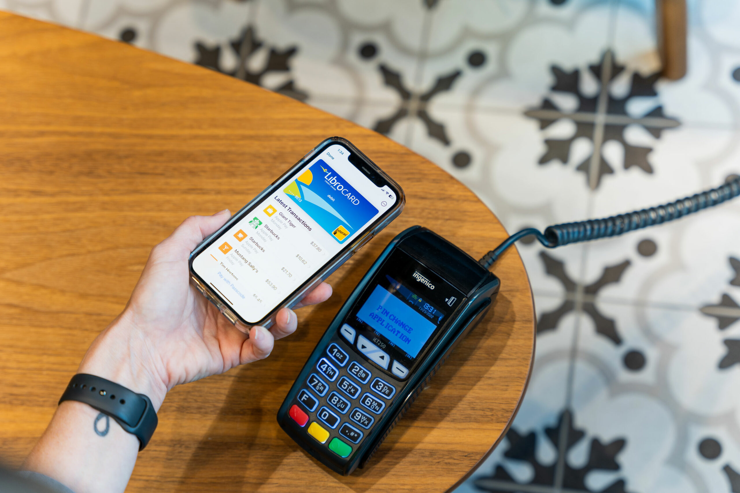 A person holds a smartphone displaying a digital wallet app near a card payment terminal on a wooden table, ready for contactless payment.