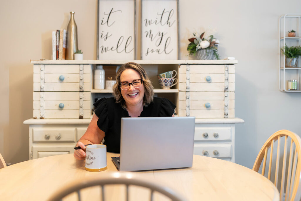 A woman sits at a round table with a laptop and a mug, smiling at the camera. She is in a cozy room with wooden furniture and wall decor in the background.
