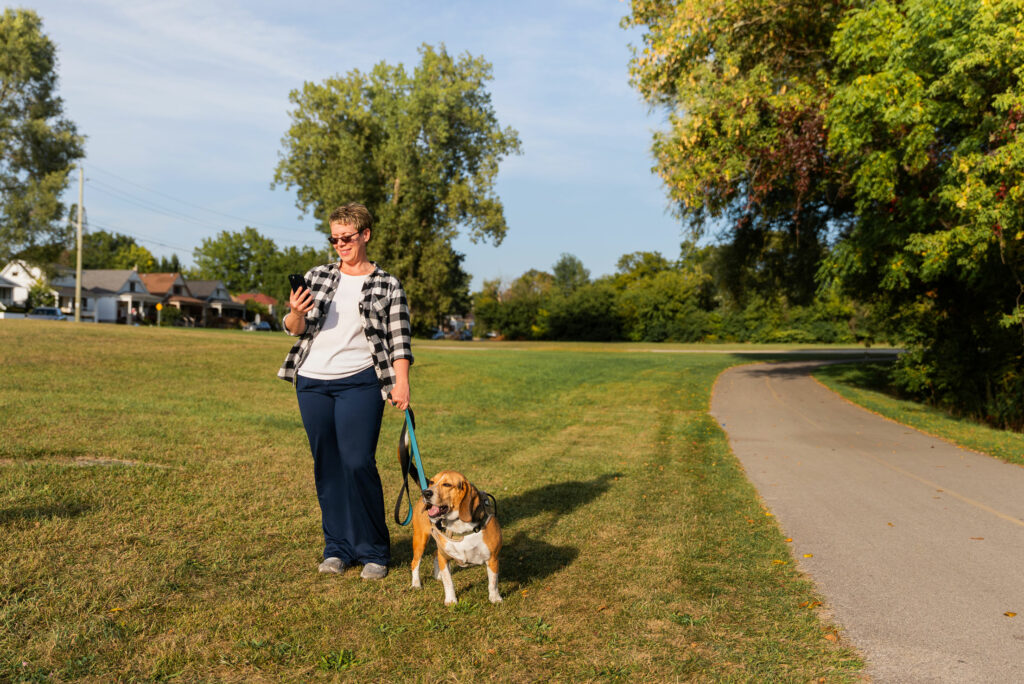 A person stands on grass beside a paved path, holding a phone and a leash attached to a brown and white dog; trees and houses are in the background.