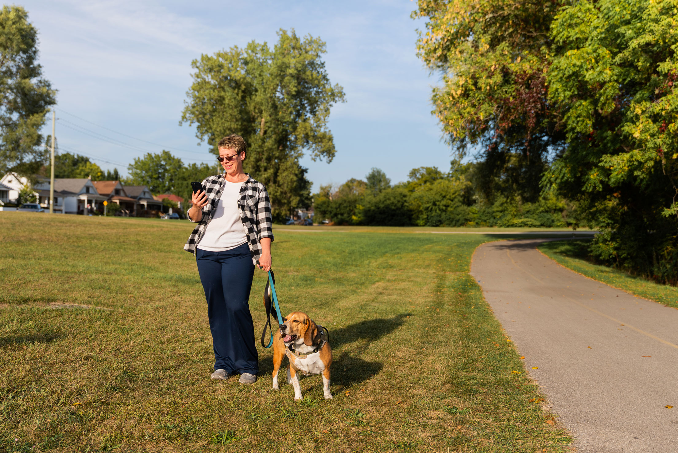A person stands on grass beside a paved path, holding a phone and a leash attached to a brown and white dog; trees and houses are in the background.