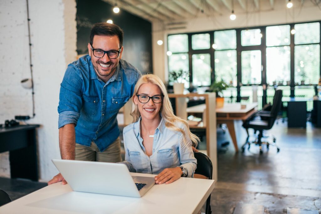 A man and a woman wearing glasses and denim shirts smile while working together at a laptop in a modern, bright office space.