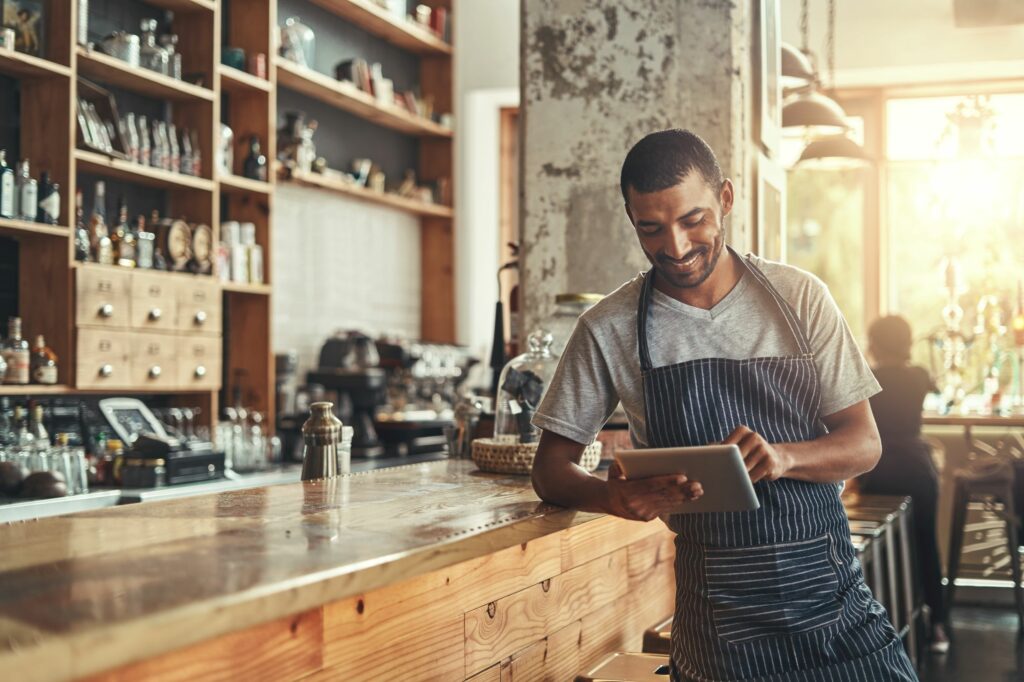 A man wearing an apron stands behind a bar counter, smiling while using a tablet in a cafe or restaurant setting.