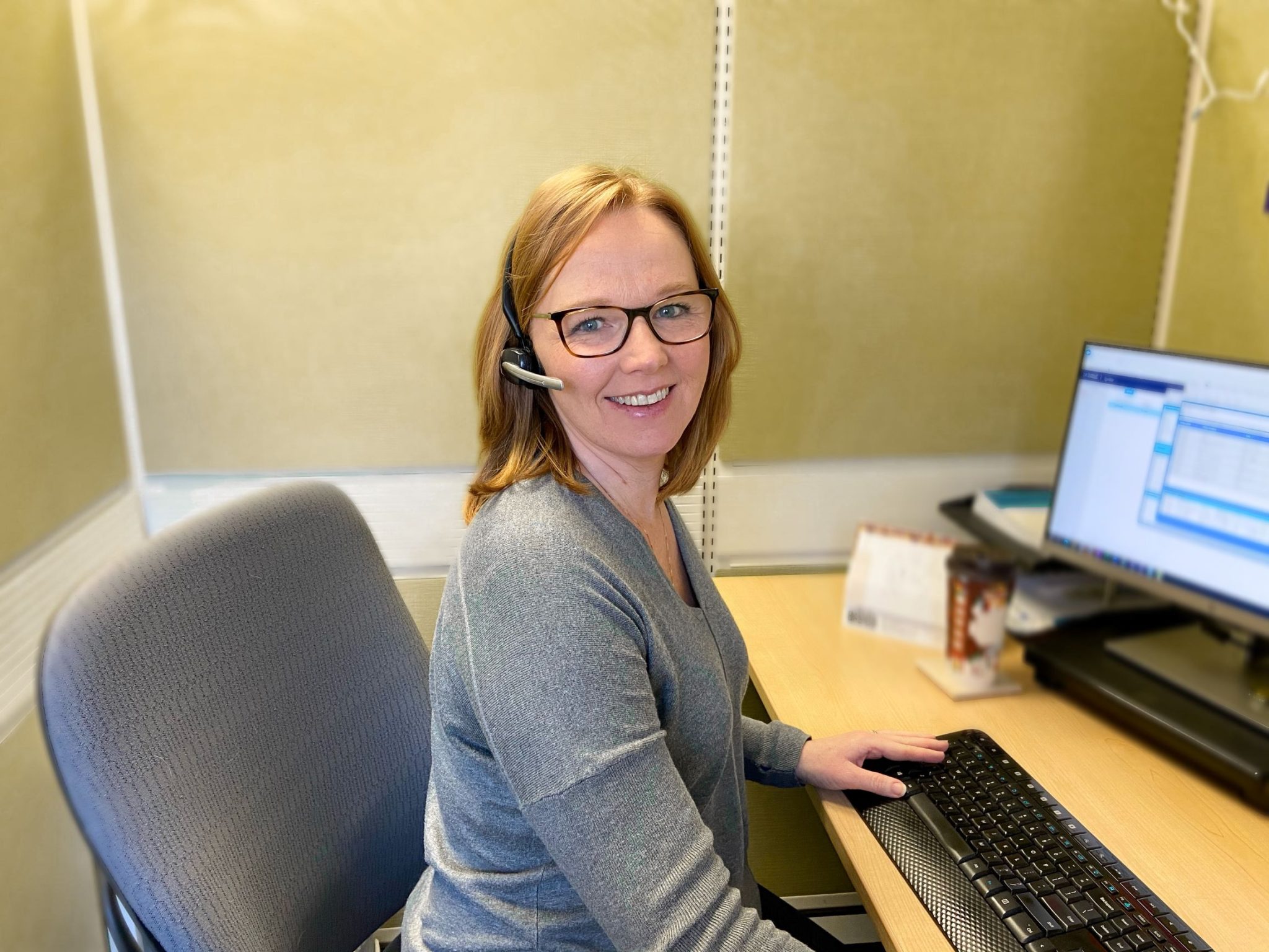 A woman wearing glasses and a headset sits at a desk with a computer, looking at the camera and smiling.