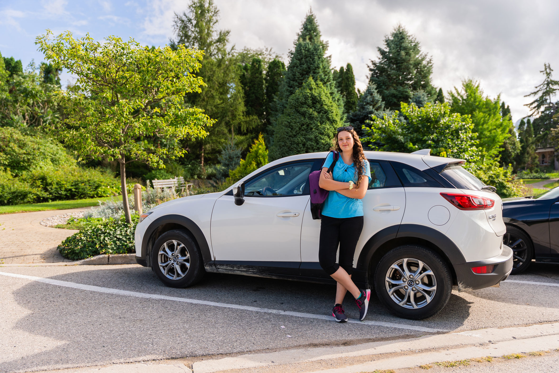 A woman in casual clothes stands with arms crossed, leaning against a white SUV parked in a sunny, tree-lined parking lot.