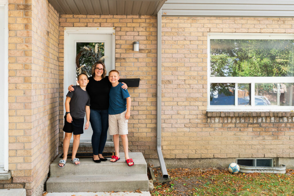 A woman stands on a small porch with her arms around two children in front of a brick house; a soccer ball lies on the grass nearby.