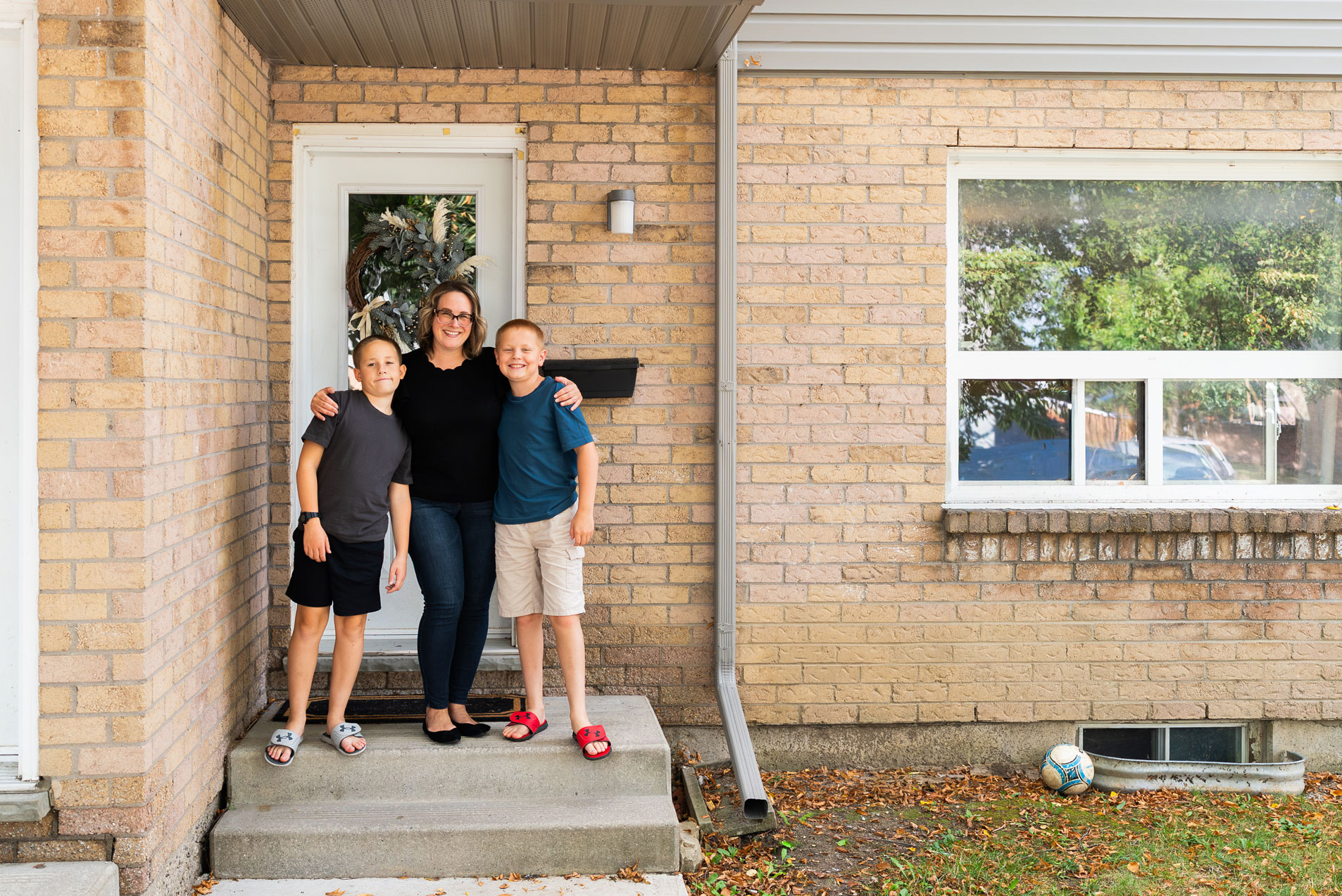 A woman stands on a small porch with her arms around two children in front of a brick house; a soccer ball lies on the grass nearby.