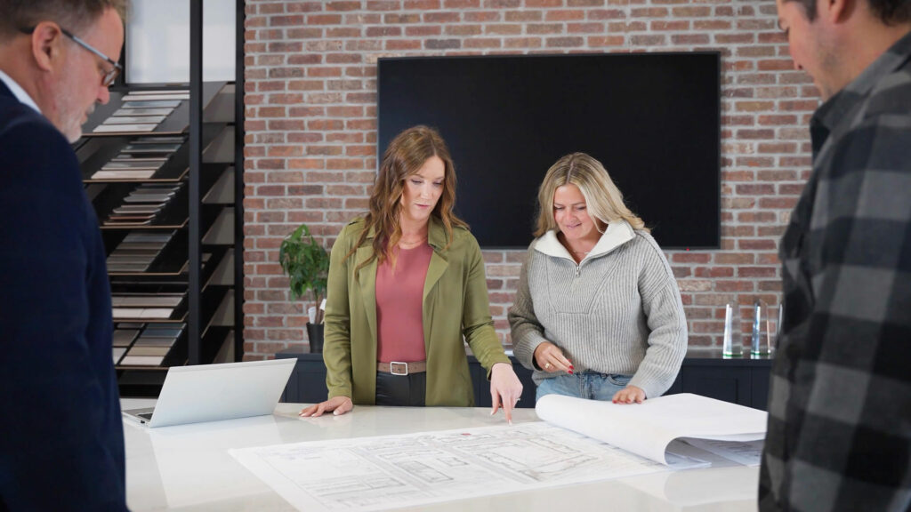 Four people stand around a table reviewing architectural blueprints in a modern office with shelves, a TV, and a laptop visible in the background.