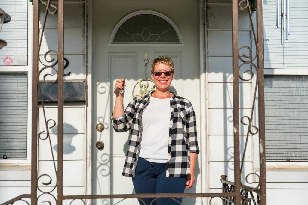 A person stands on a porch holding a key and smiling in front of a white door, wearing sunglasses, a plaid shirt, and dark pants.