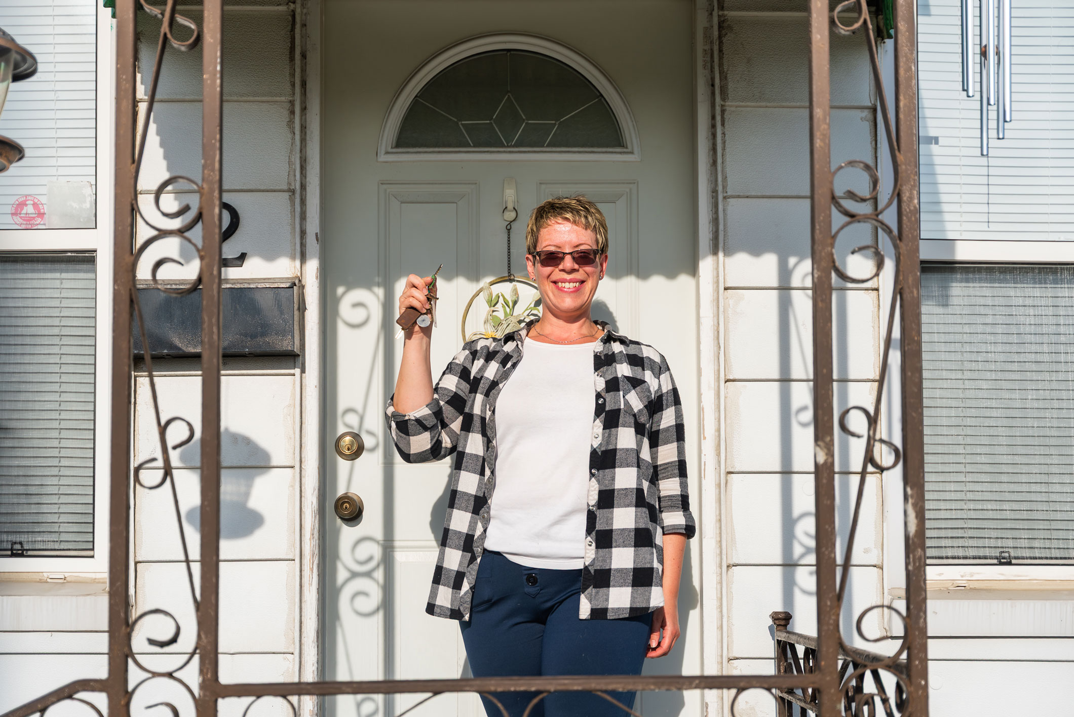 A person stands on a porch holding a key and smiling in front of a white door, wearing sunglasses, a plaid shirt, and dark pants.