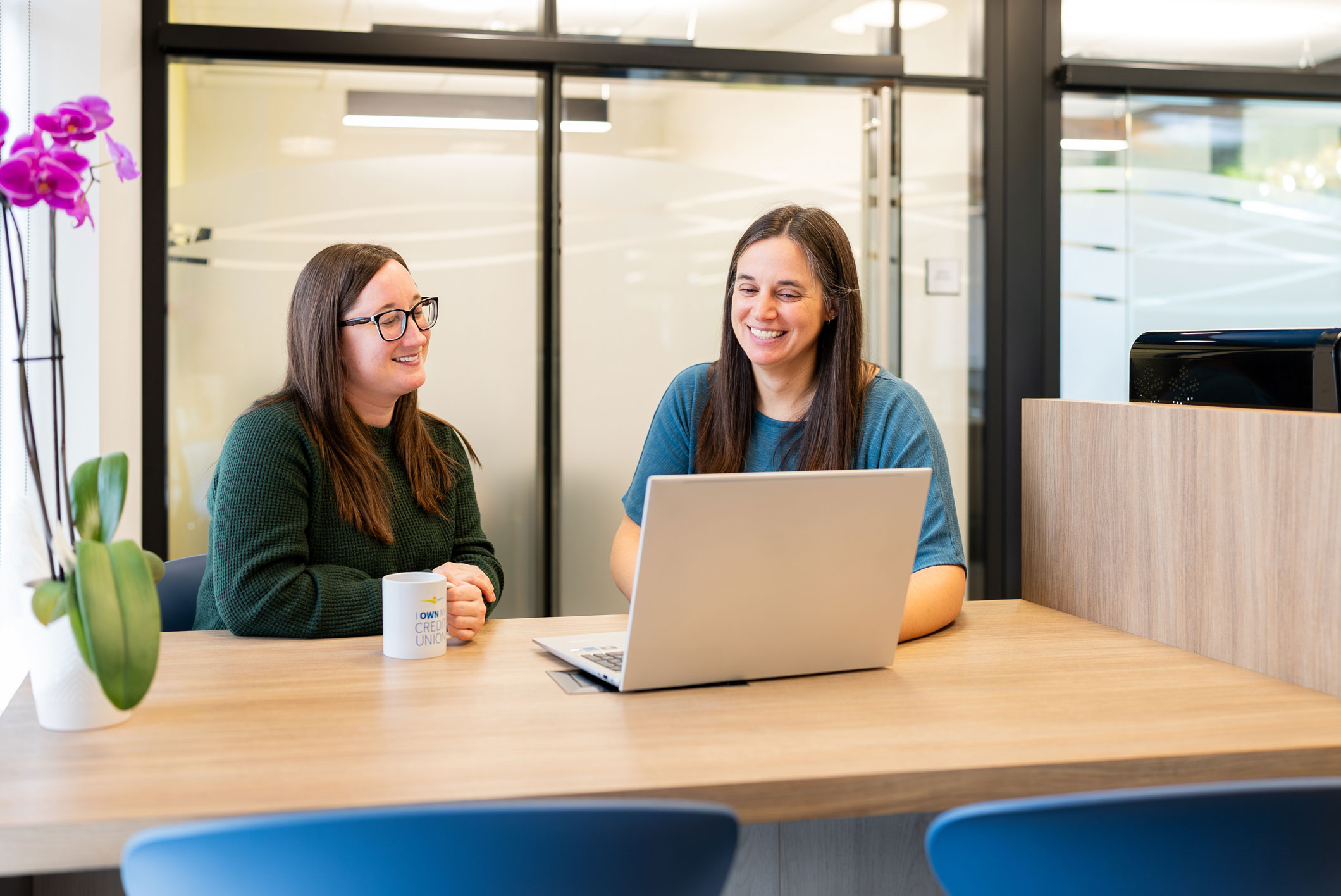 Two women sit at a desk with a laptop, smiling and talking. One holds a coffee mug. An orchid is on the left, with glass office walls in the background.