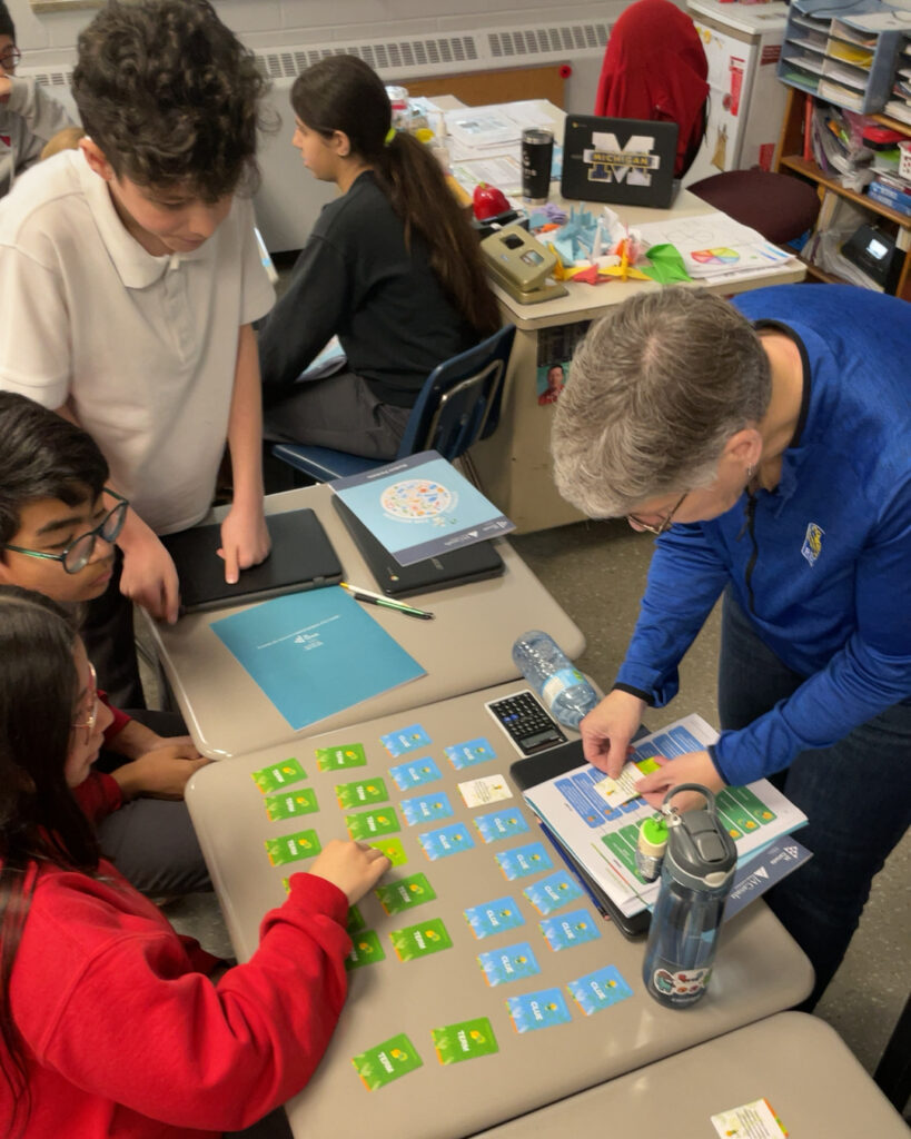 A teacher assists students working with colorful cards on a classroom desk. Two students are seated while two others and the teacher stand, engaging with the activity.