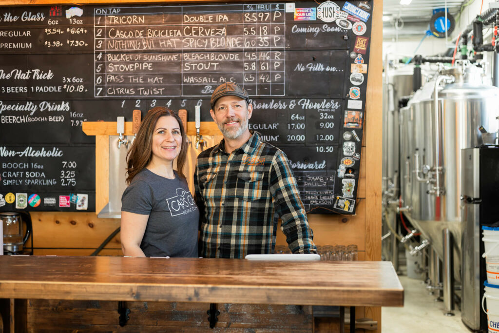 Two people stand behind a wooden bar in a brewery, smiling at the camera, with a chalkboard menu and stainless steel brewing tanks in the background.