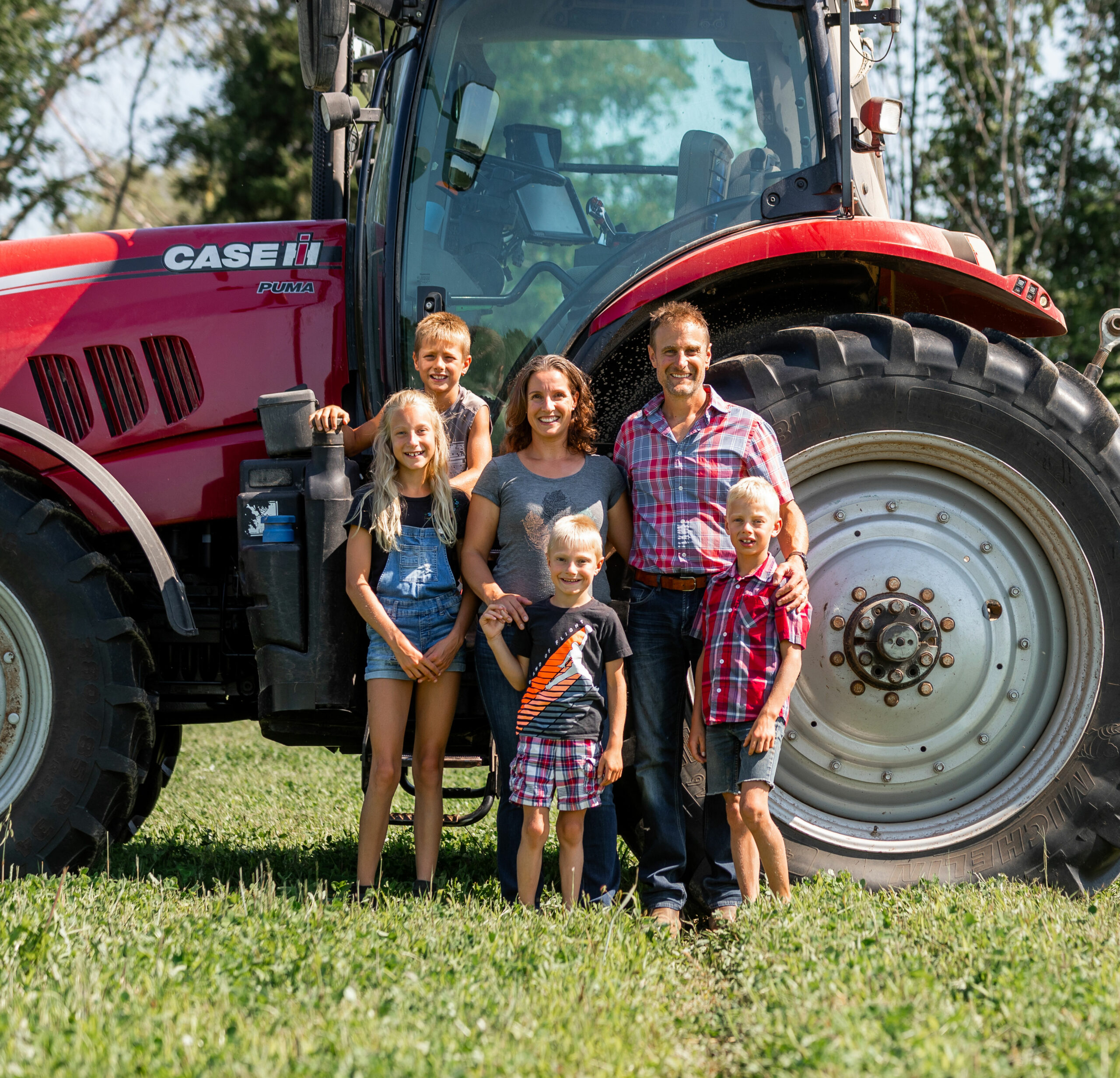 A family of five stands smiling in front of a large red Case IH tractor on a grassy field, with trees visible in the background.
