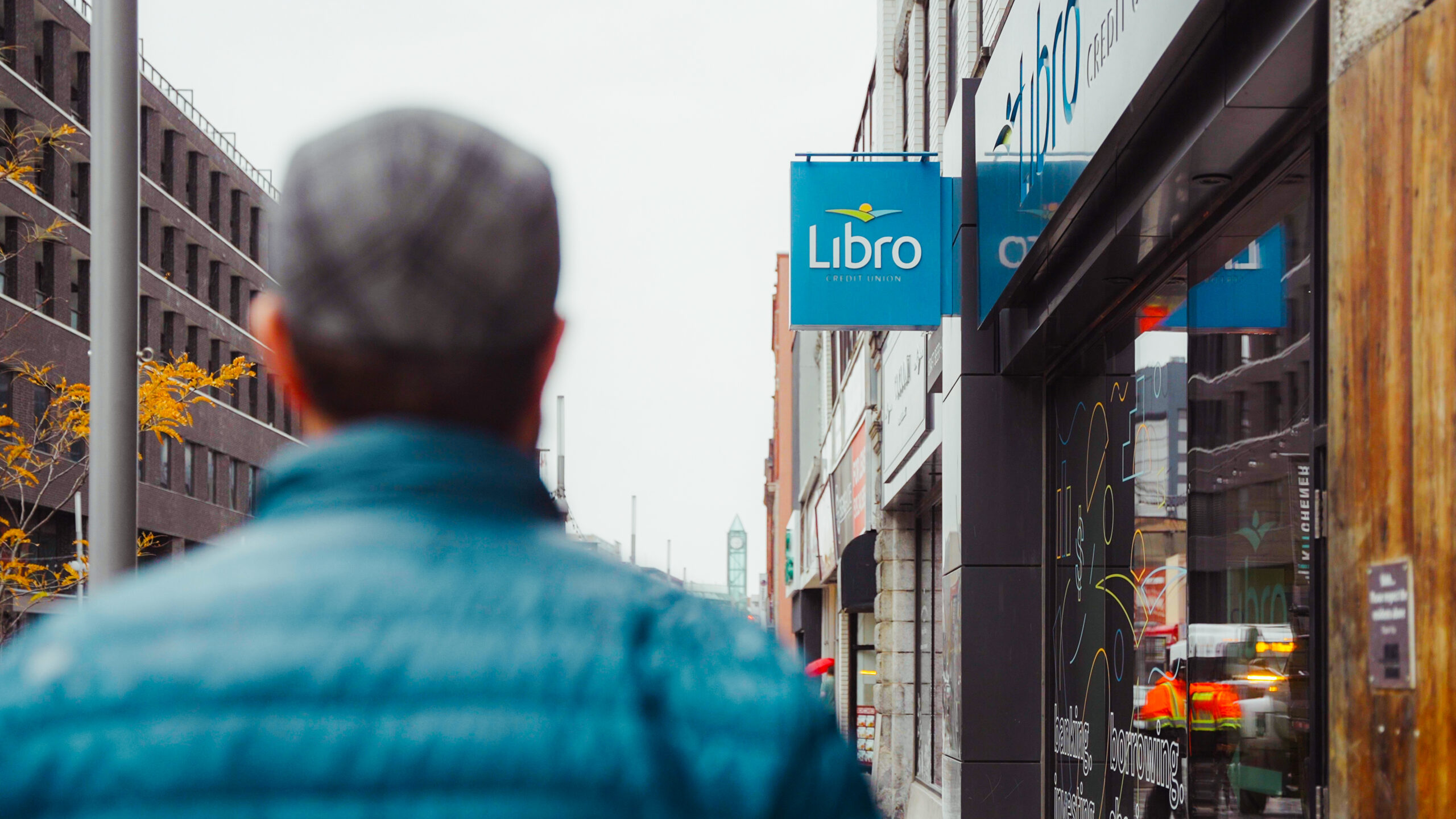 A person in a blue jacket walks down a city sidewalk toward a building with a blue "Libro" sign.