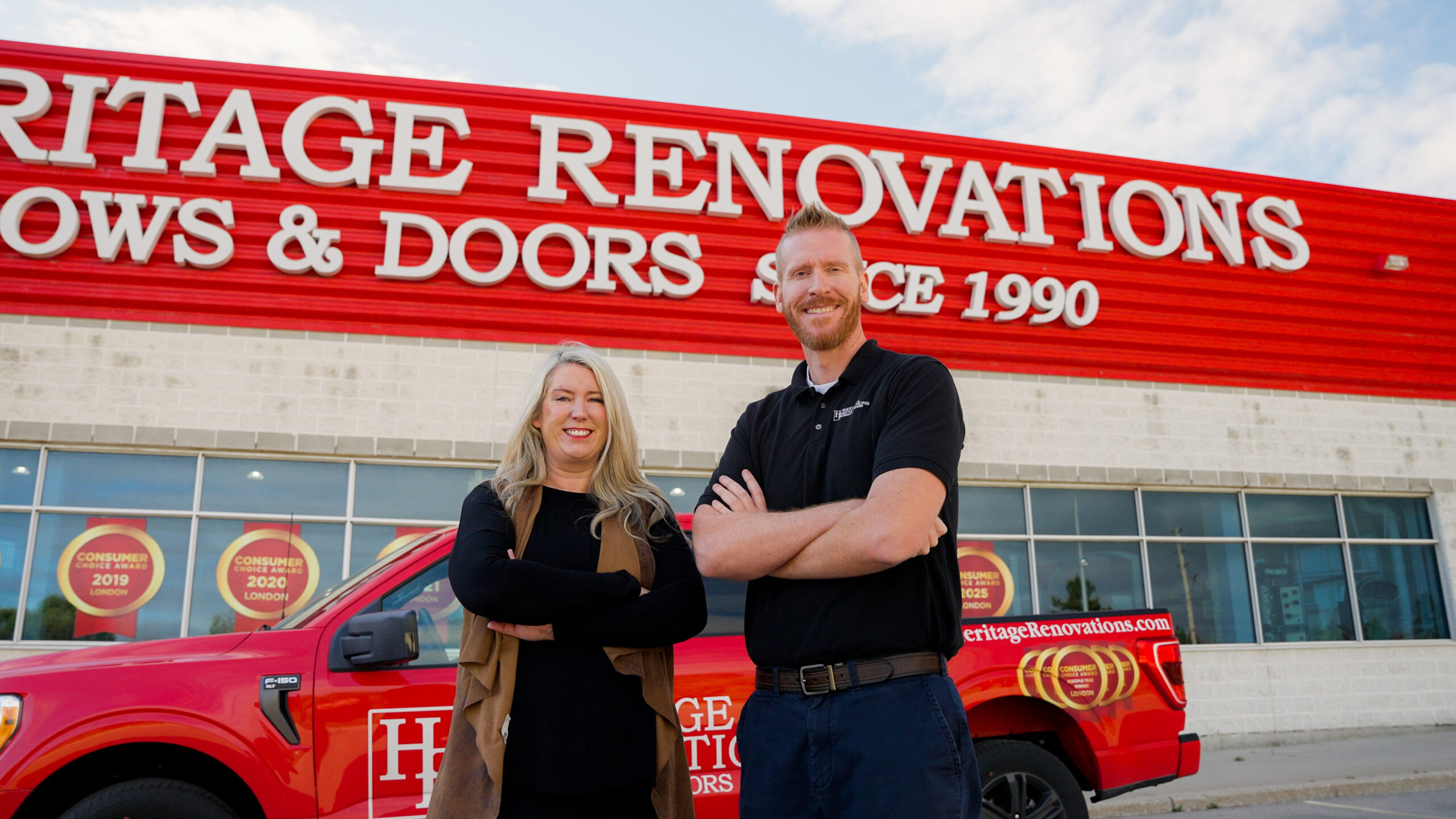 Two people stand smiling with arms crossed in front of a red truck and a building labeled “Heritage Renovations Windows & Doors - Since 1990.”.