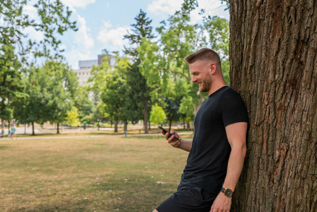 Libro Owner, A.J., looking at his phone in a park and leaning against a tree trunk.