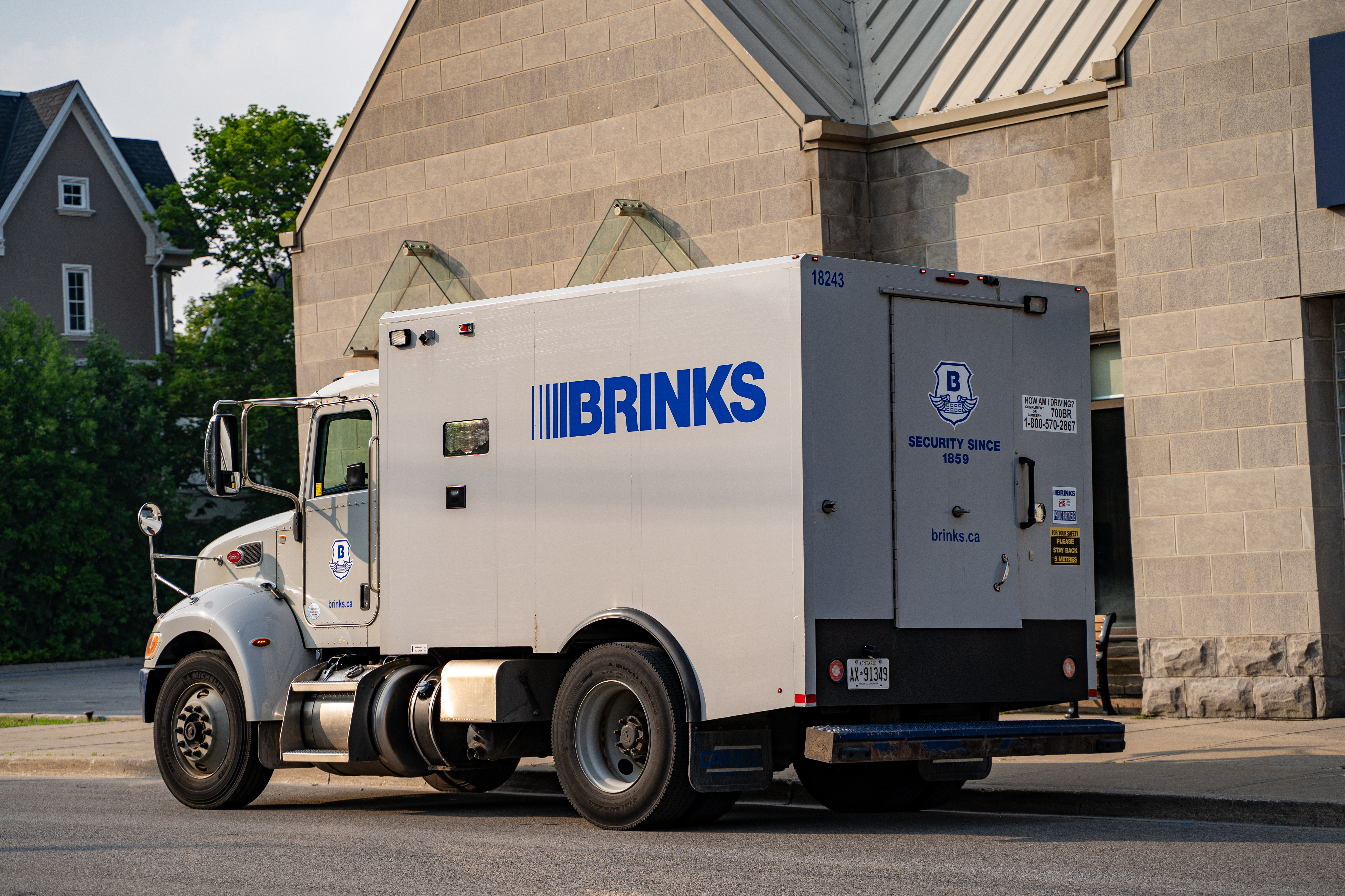 A Brinks armored truck is parked on the street beside a gray stone building on a sunny day.