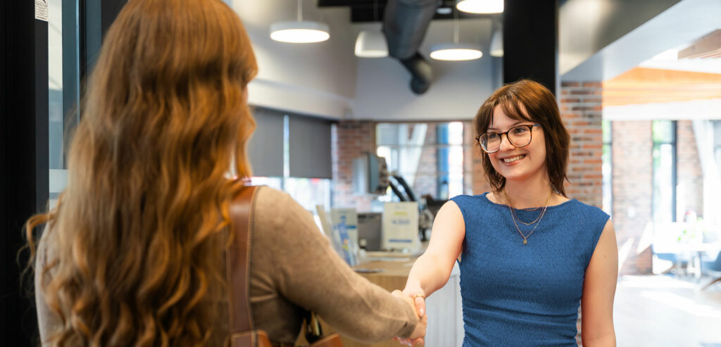 Two women are shaking hands in a modern office setting, with one woman facing the camera and smiling.