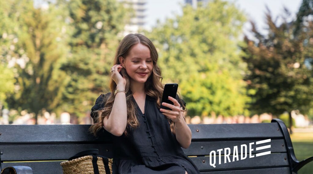A woman sits on a park bench, smiling while looking at her phone, with a woven bag beside her and the QTRADE logo in the corner.
