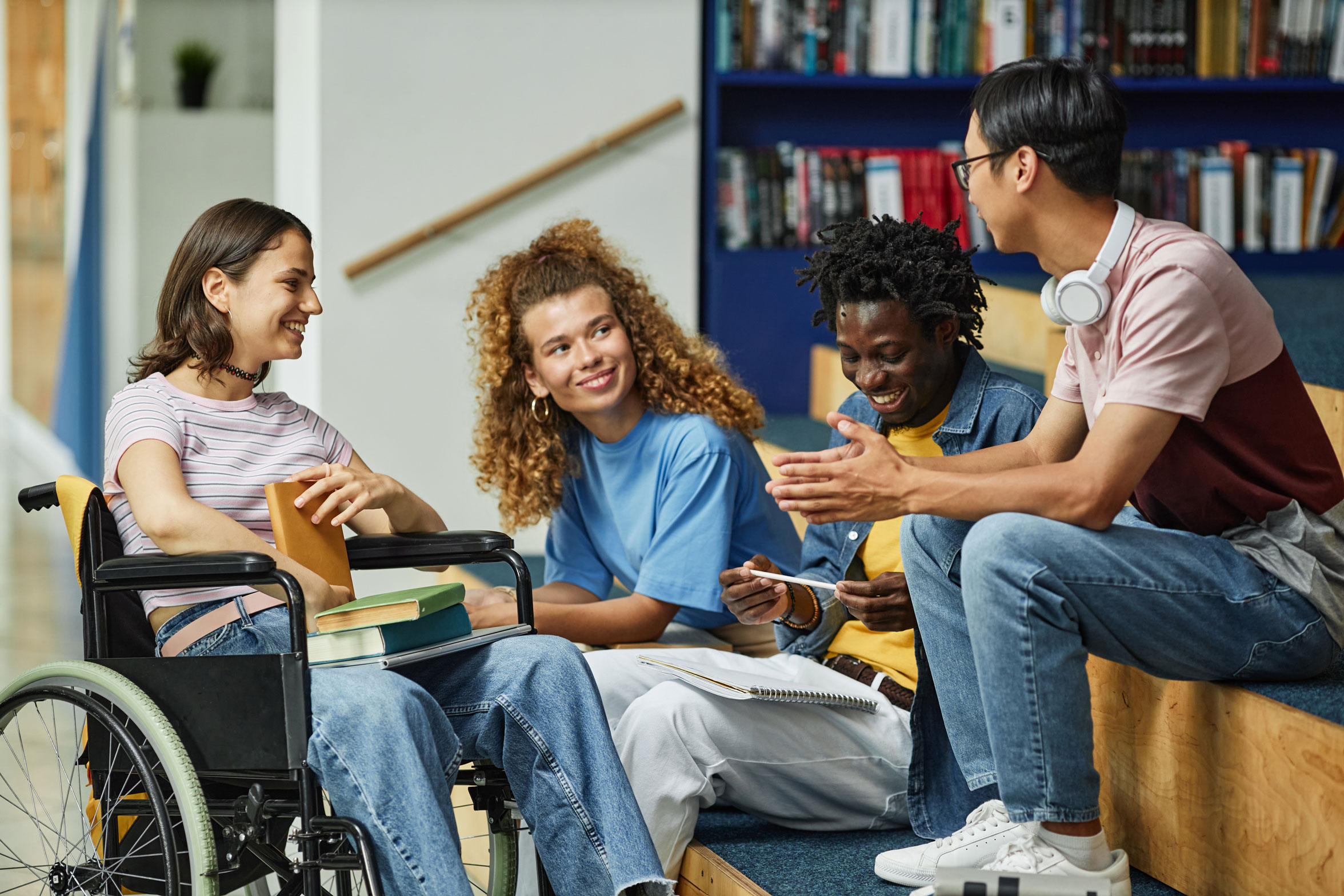 Four young adults, one using a wheelchair, sit and talk together in a library, holding books and notebooks.