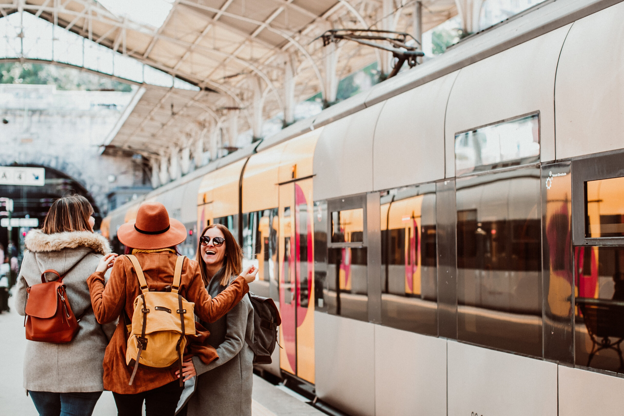 Three people with backpacks stand on a train platform, smiling and talking, next to a modern train under a covered station.