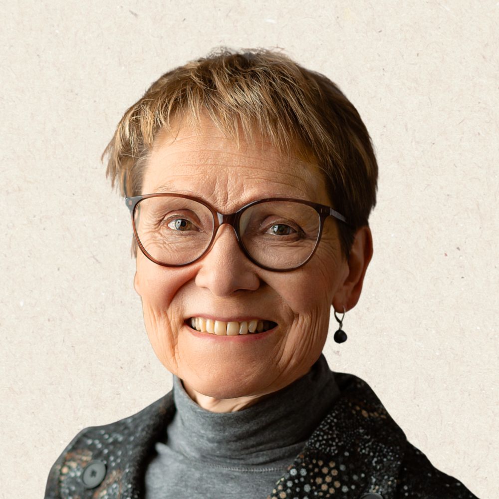 Older woman with short hair, glasses, and earrings, smiling at the camera while wearing a turtleneck and patterned jacket against a neutral background.