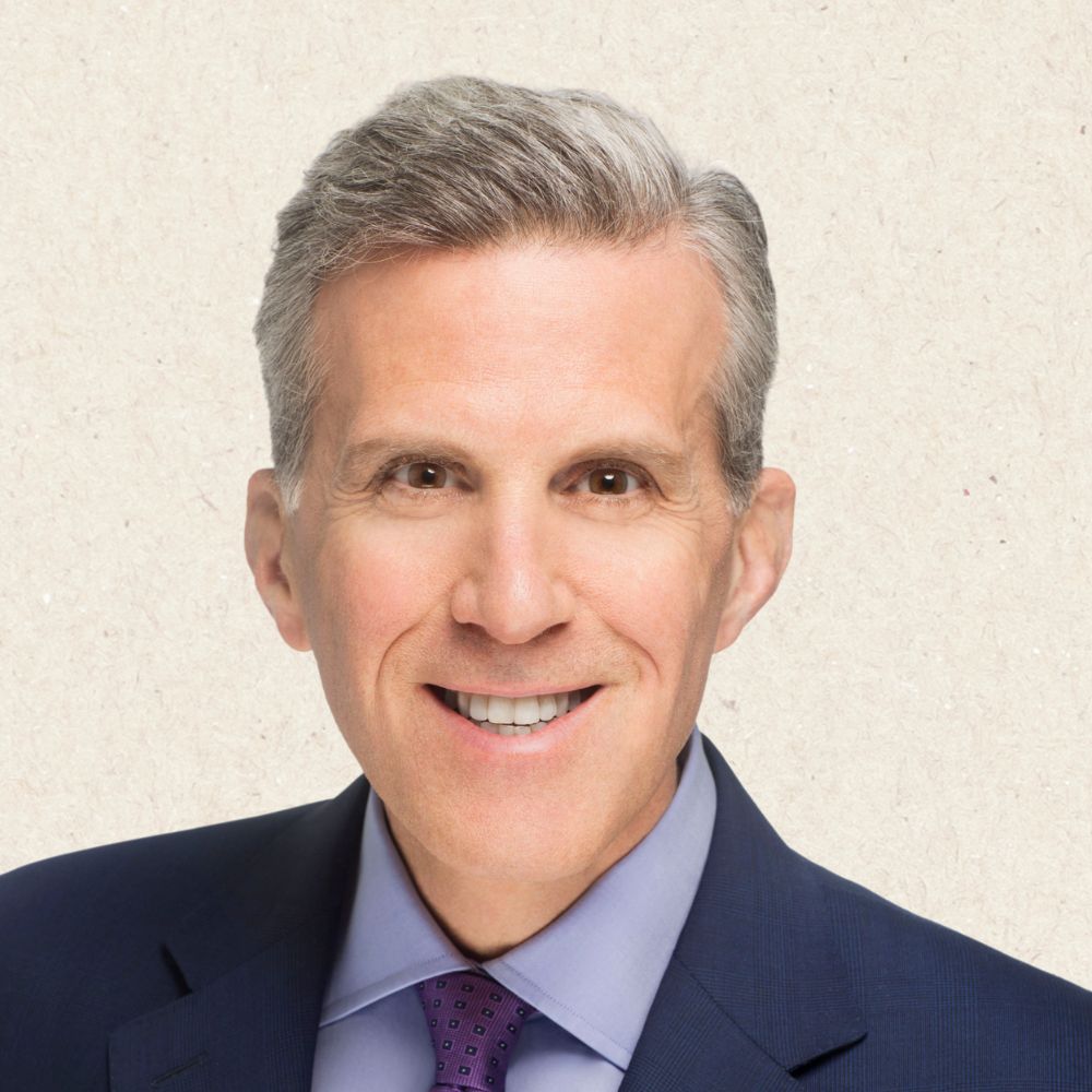 Smiling middle-aged man with short gray hair wearing a dark suit, light blue shirt, and purple patterned tie, posed against a plain light background.