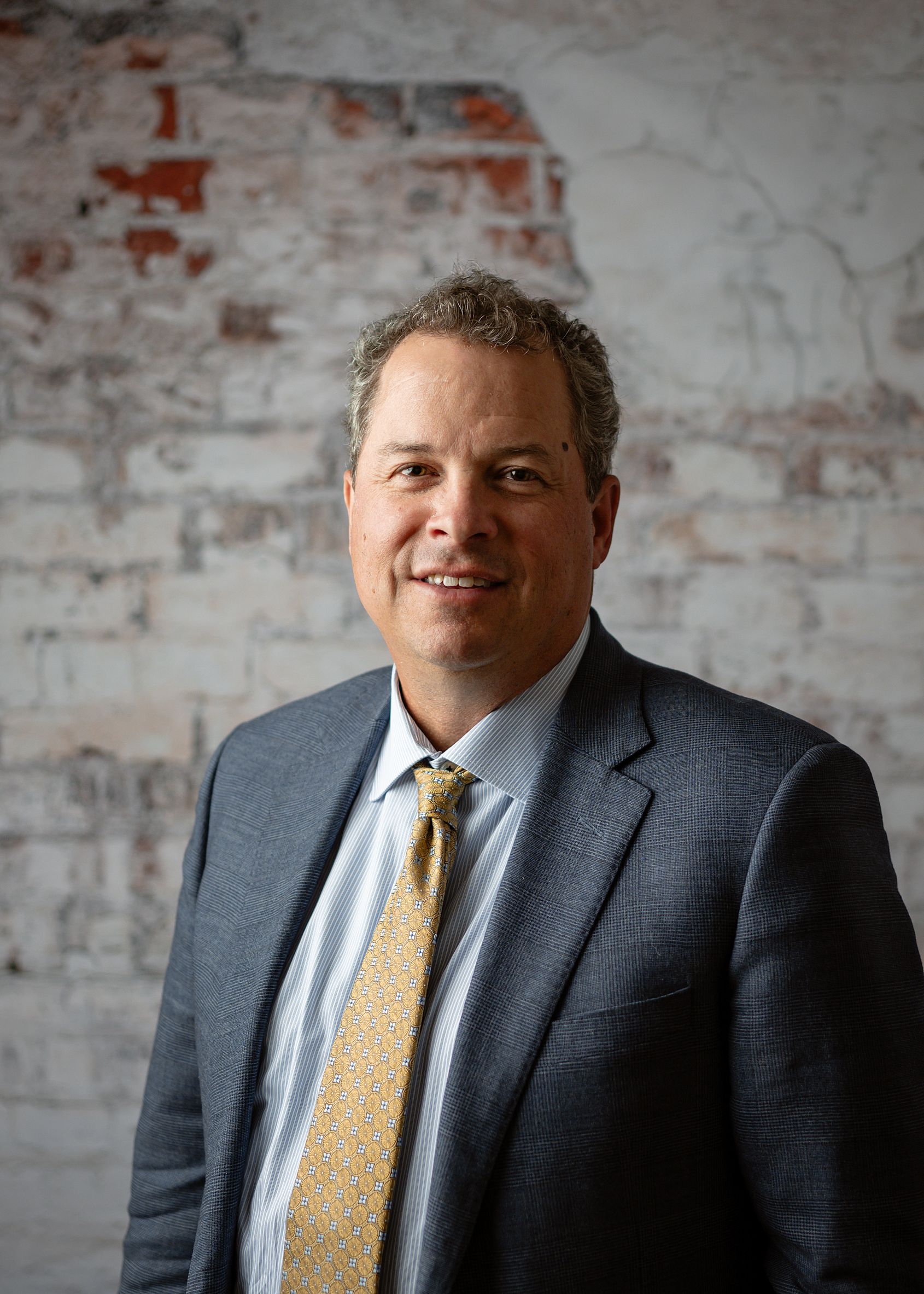 A man in a suit and tie stands in front of a white brick wall, looking at the camera with a neutral expression.