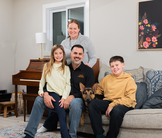 A family of four and a dog pose together in a living room, with a piano, floral painting, and window in the background—bank local and support local like Libro, offering banking and financial services in Ontario.