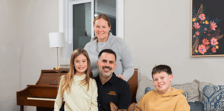 A woman, man, two children, and a dog pose together in a living room with a piano, window, and floral painting in the background.