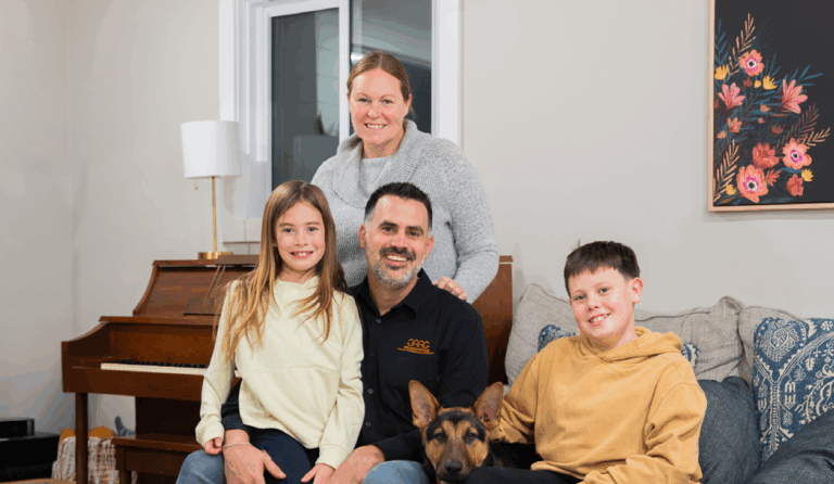 A woman, man, two children, and a dog pose together in a living room with a piano, window, and floral painting in the background.