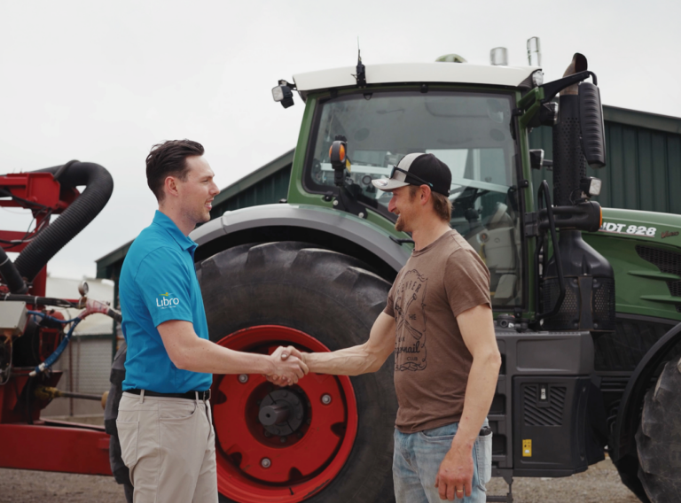 Two men shake hands in front of a large green tractor and farm equipment, with a barn visible in the background.