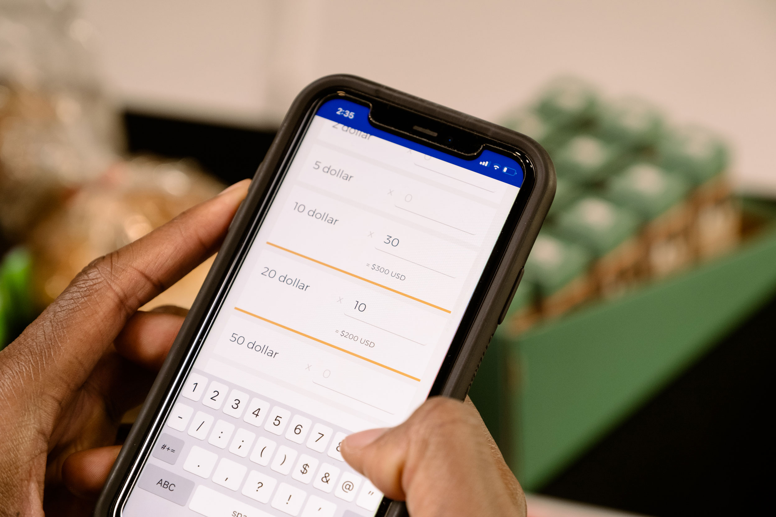 A person uses a smartphone to enter dollar amounts in an app, with food items and a green carton visible in the blurred background.