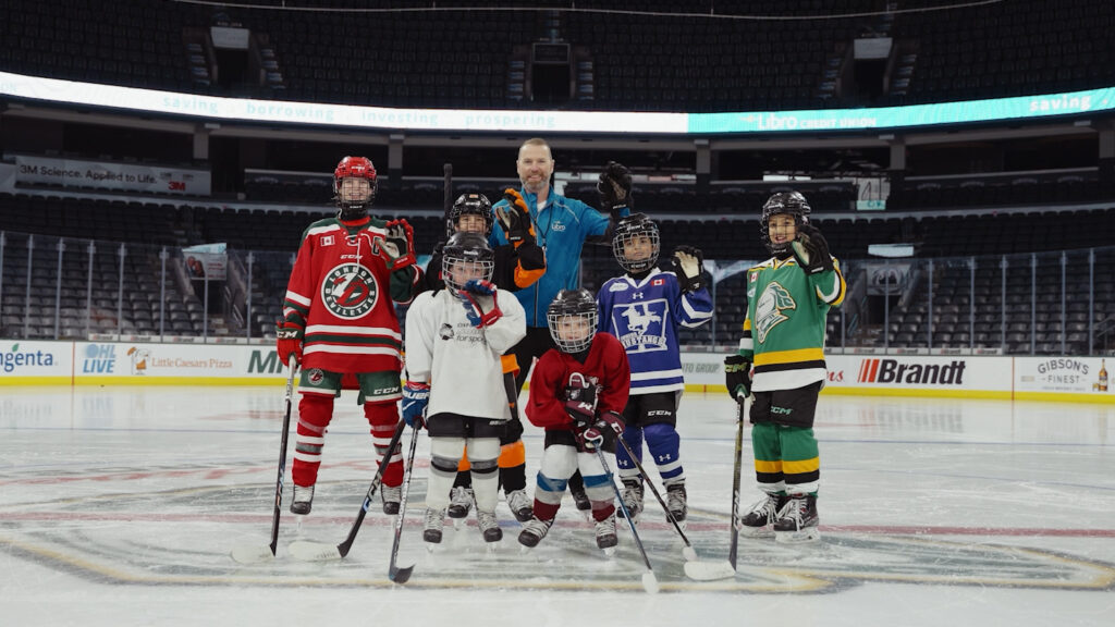 A group of six children in hockey uniforms and one adult stand on an ice rink, posing for a photo with hockey sticks.