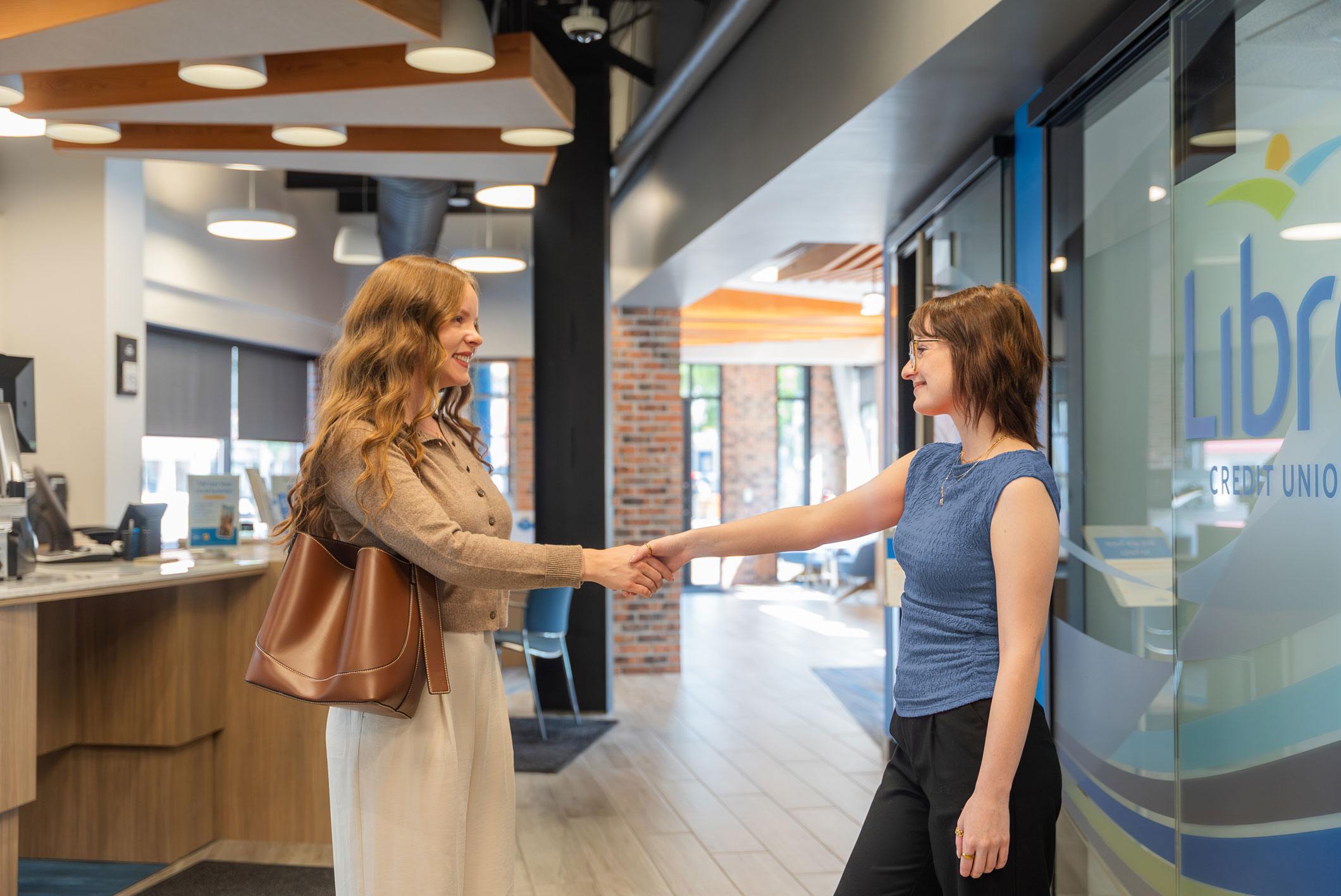 Two women are shaking hands in a modern office lobby near a glass door with the words 