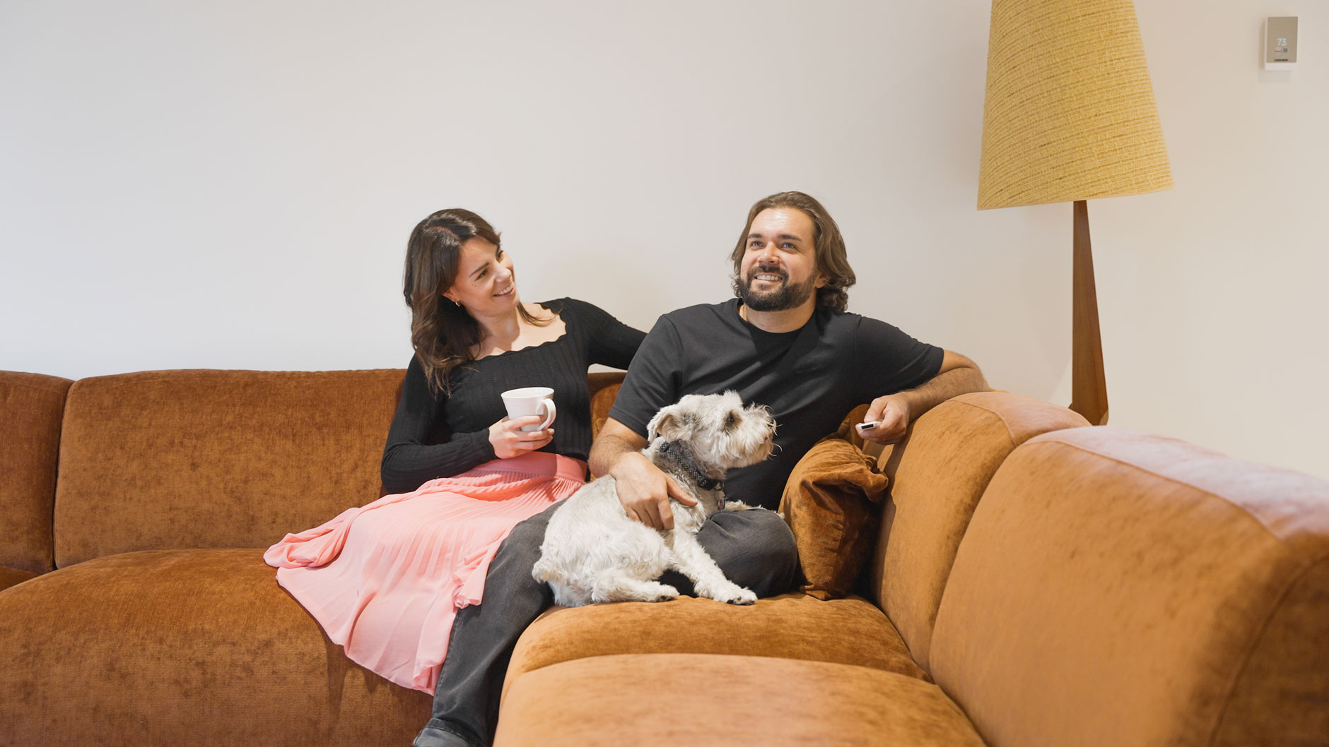 A woman and man sit on a brown sofa with a small dog on the man's lap; the woman holds a mug and both appear relaxed.