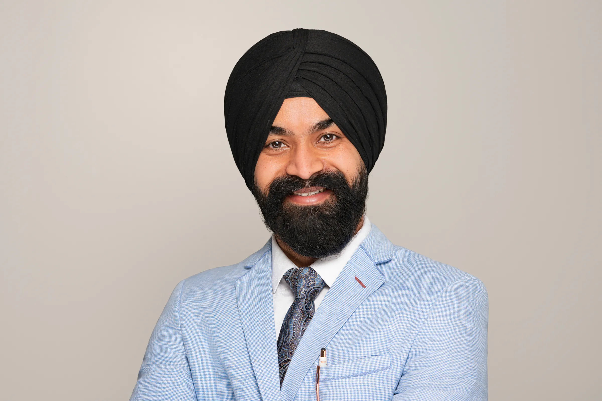 A man wearing a black turban and light blue suit jacket stands smiling with his arms crossed against a plain background.