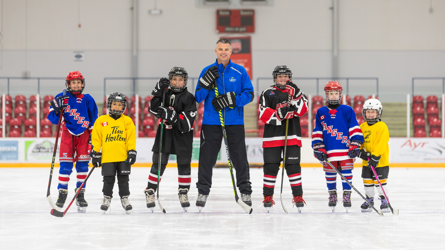A group of six children in hockey gear and one adult coach stand on an indoor ice rink, posing for a photo with hockey sticks.