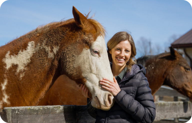 A woman wearing a black jacket stands outdoors next to a brown and white horse, smiling while petting its face, with another horse visible in the background.