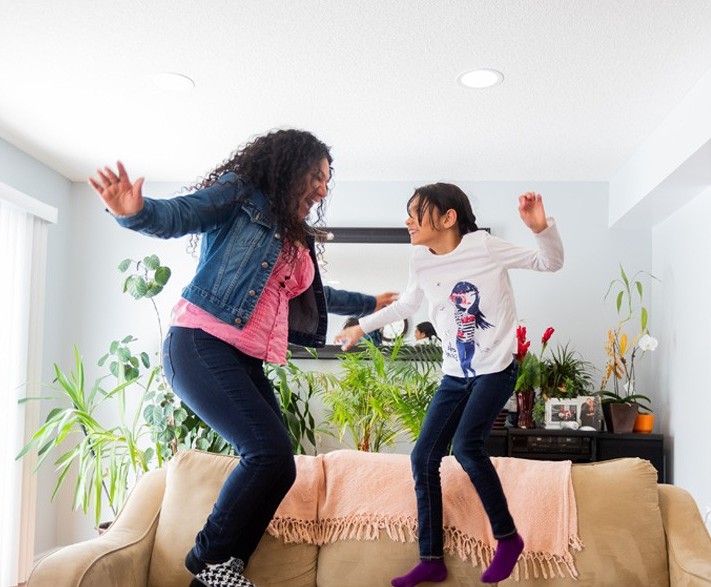 Two people, an adult and a child, are jumping on a beige couch in a living room with plants and a mirror in the background.
