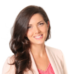 A woman with long brown hair wearing a light pink blazer and a coral top smiles at the camera against a white background.