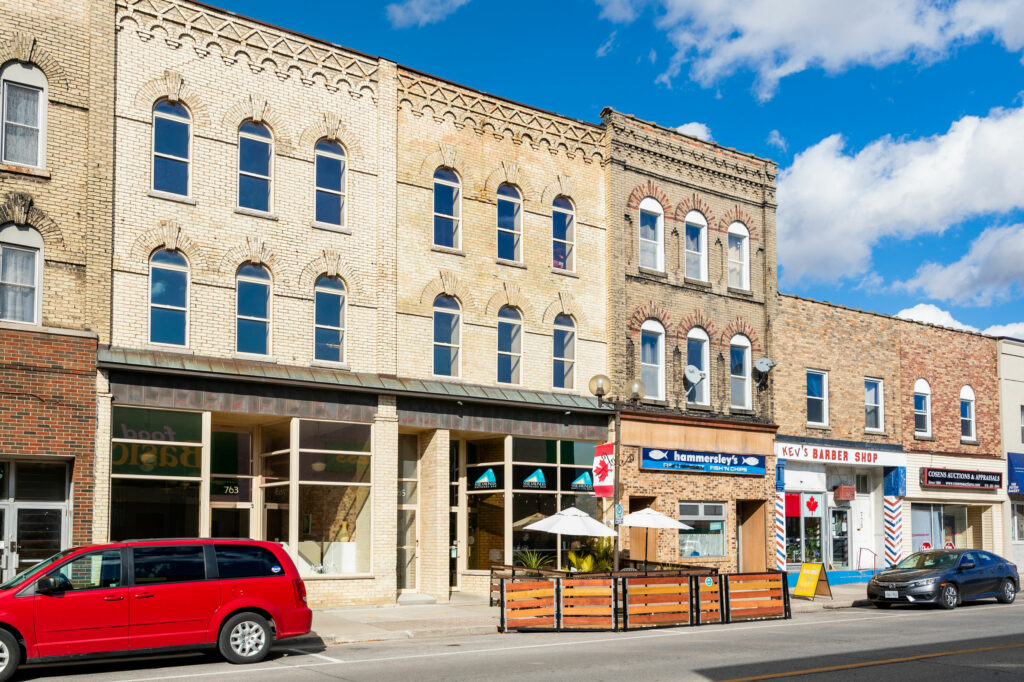 A row of brick commercial buildings with shops, outdoor seating, and parked cars along a sunny street under a partly cloudy sky.