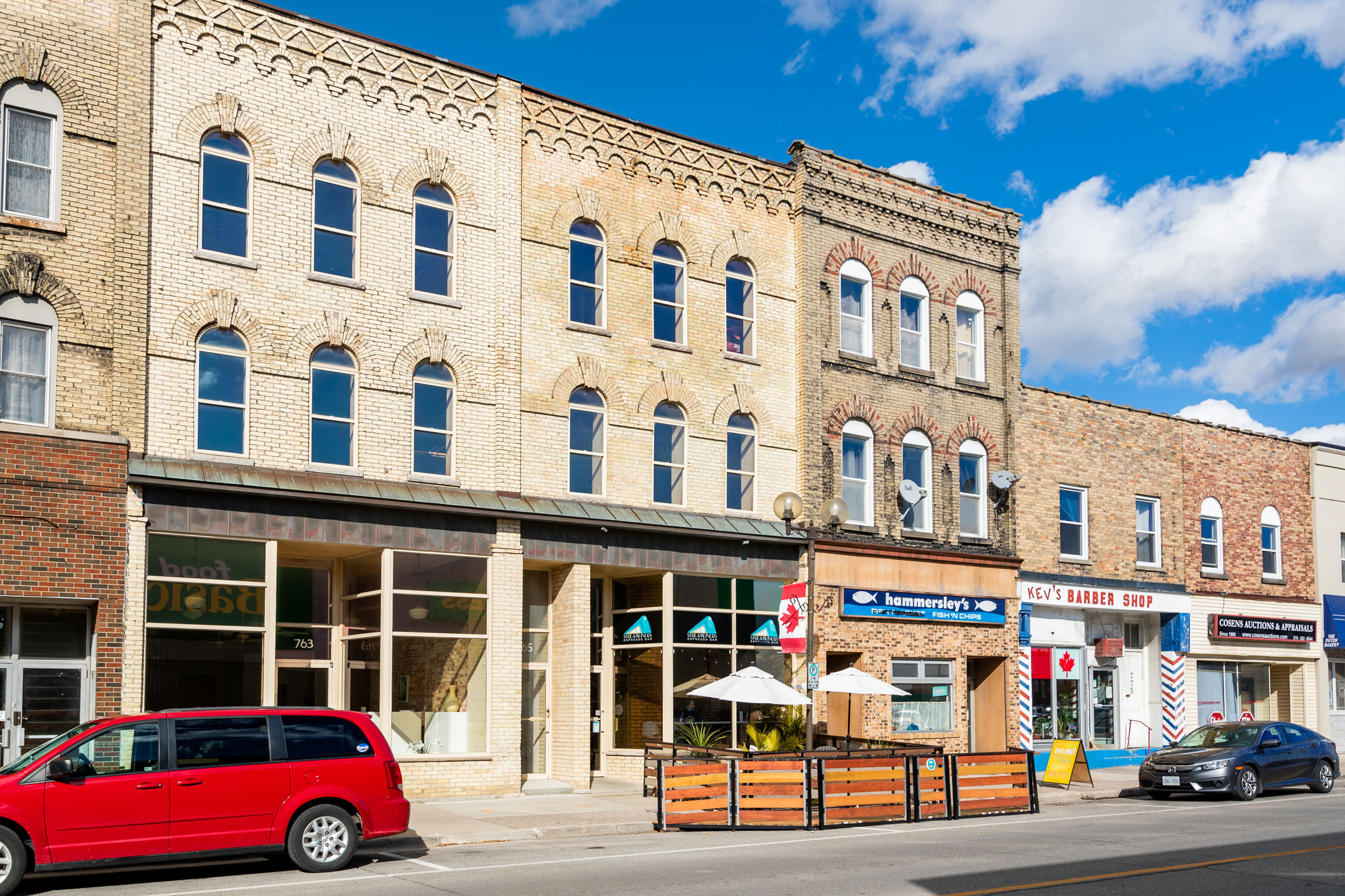 A row of brick commercial buildings with shops, outdoor seating, and parked cars along a sunny street under a partly cloudy sky.