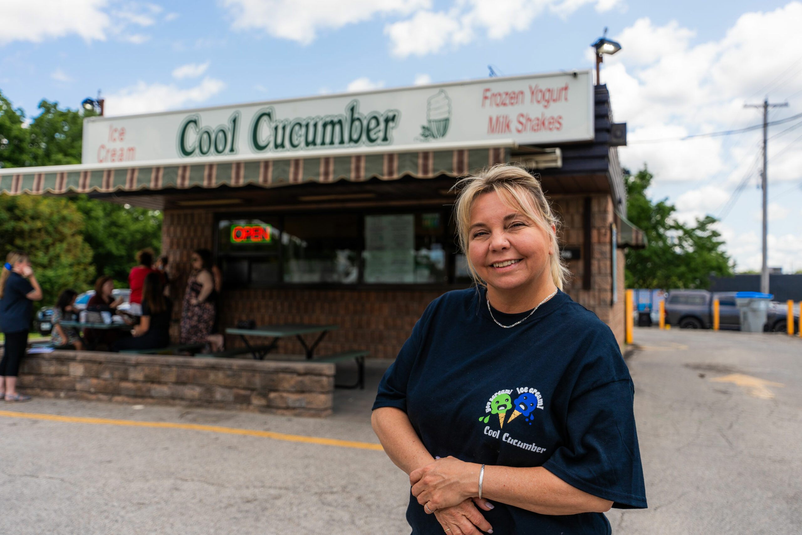 A woman stands in front of an ice cream shop called "Cool Cucumber" with an open sign; several people are lined up by the window.