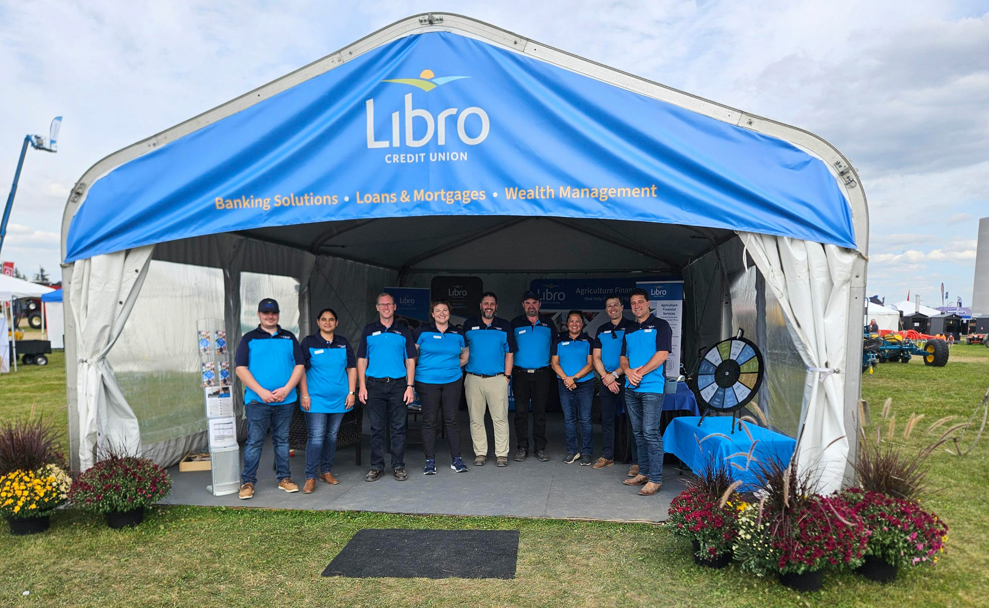 Nine people wearing blue shirts stand in front of a Libro Credit Union tent at an outdoor event, surrounded by potted flowers and informational displays.
